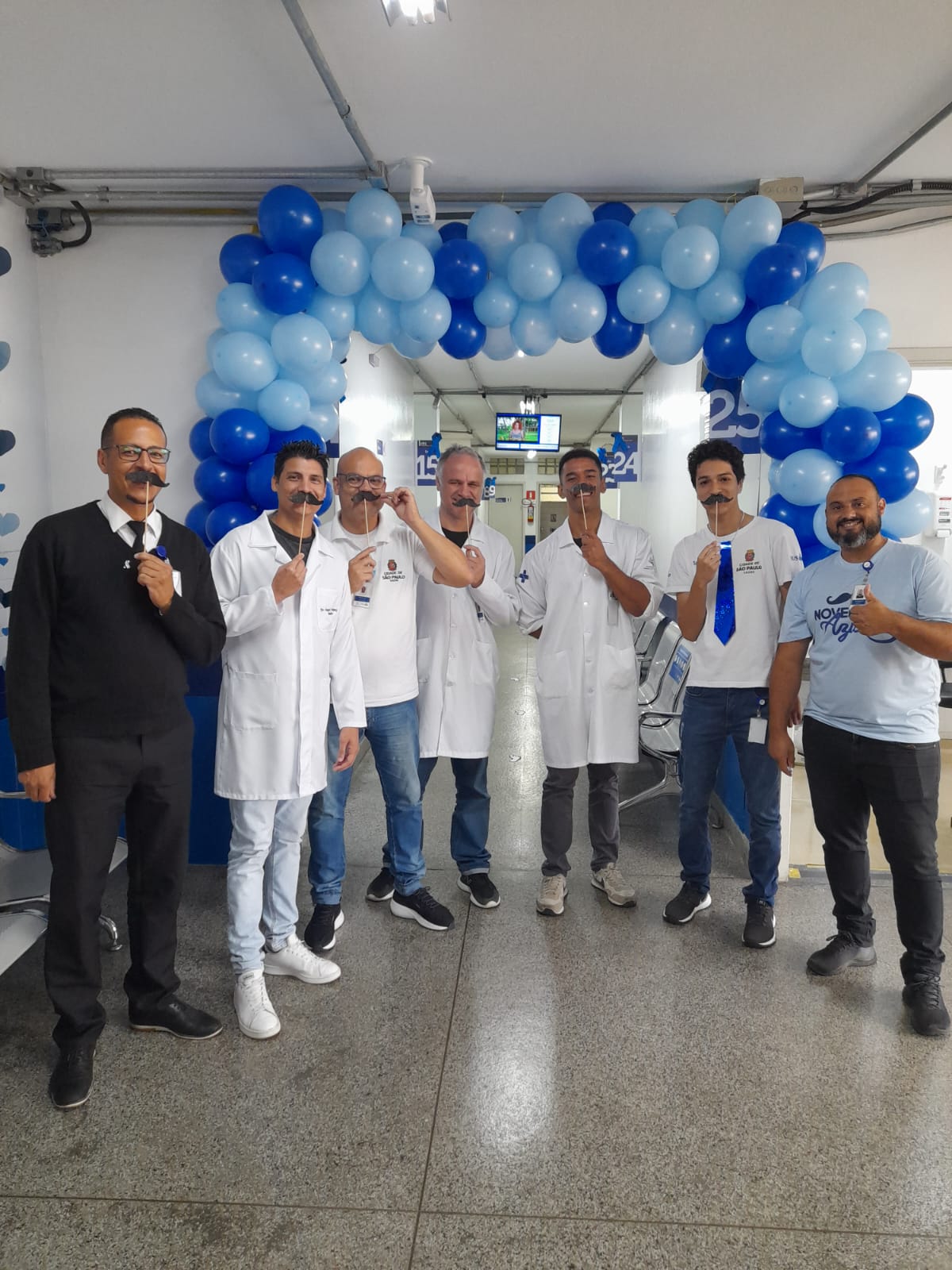 A foto mostra um grupo de sete profissionais da saúde, todos homens, segurando um bigode falso em frente ao rosto; eles estão na parte interna de uma unidade básica de saúde, junto a um arco feito com balões azuis